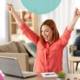 Woman sat at desk with her arms in the air whilst smiling at a laptop screen